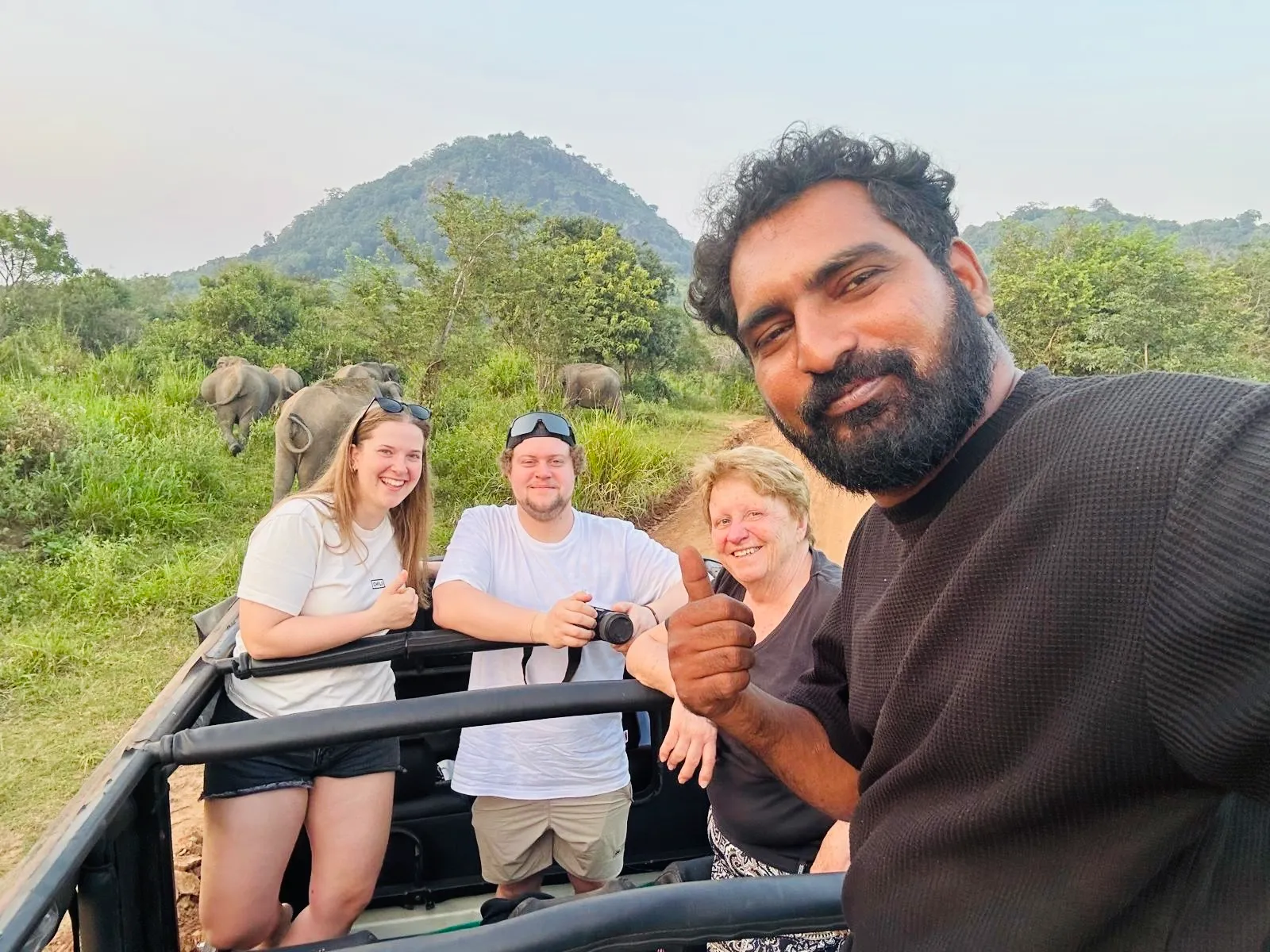 Smiling guests and their driver in an open jeep with elephants behind, on a Habarana elephant safari